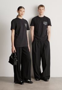 Czarne t-shirty z małym nadrukiem na lewej stronie klatki piersiowej, w połączeniu z luźnymi czarnymi spodniami. Modelka trzyma czarną torebkę.