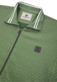 Chaqueta verde con cremallera y un patrón texturizado, cuello acanalado con rayas blancas y un parche negro con el logotipo en la parte frente.