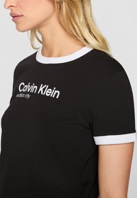 Juodas medvilninis marškinėliai su balta šonkauline apykakle ir rankogaliais. Ant priekio yra tekstas "Calvin Klein endless city" baltos spalvos.