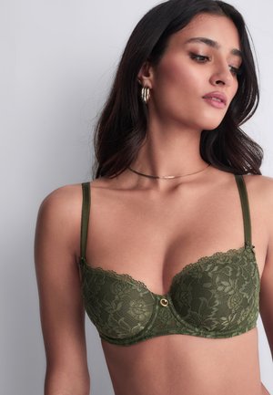 Femme aux cheveux bruns ondulés, portant des boucles d'oreilles créoles dorées et un collier fin, portant un soutien-gorge en dentelle vert olive à motif floral.