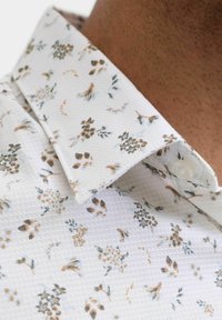 Valkoinen kuvioitu kauluspaita, jossa on teksturoitu kangas. Kukkakuvio vaimeissa väreissä ja nappiyksityiskohta kauluksessa.