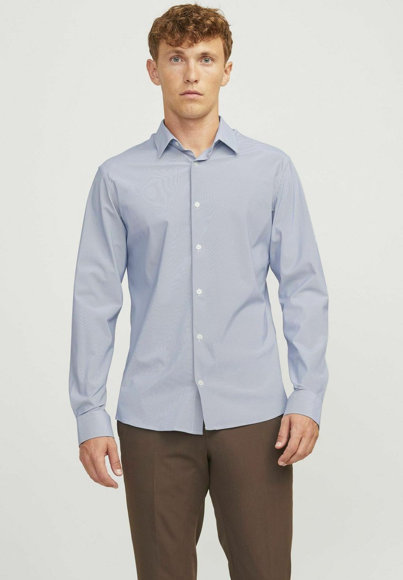 Camicia button-up azzurro chiaro con una sottile texture, maniche lunghe e colletto a punta, abbinata a pantaloni marroni. Bottoni bianchi come dettagli.
