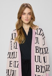 Blondynka z luźnymi falującymi włosami nosi czarną bluzkę i różowy kardigan z czarnymi wyraźnymi literami na beżowym tle.