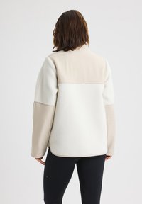 Beżowo-kremowa kurtka z polarem na korpusie i gładką tkaniną na ramionach, z lekko zaokrąglonym dołem i bocznymi rozcięciami.