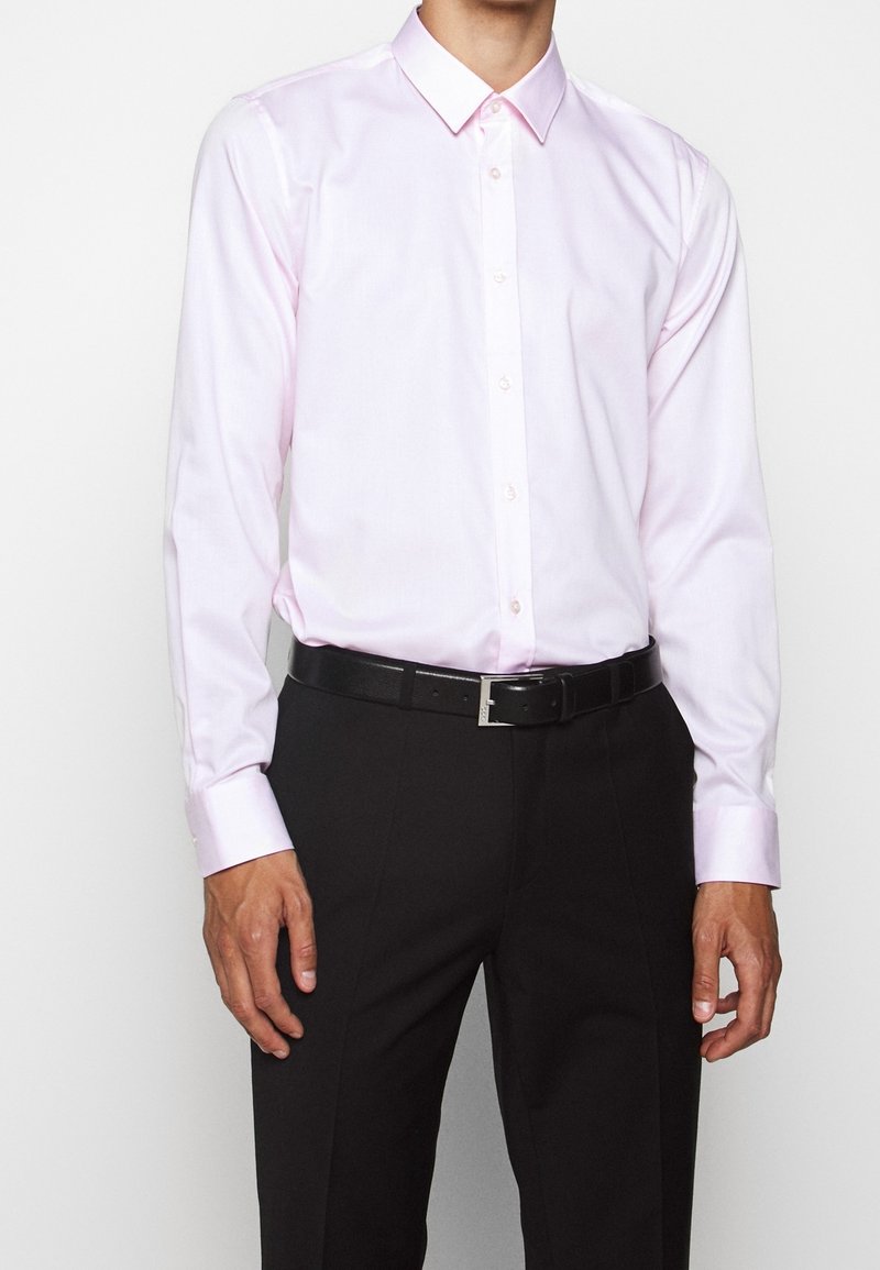 Jasnoróżowa koszula z klasycznym kołnierzykiem, długimi rękawami i zapięciem na guziki; zestawiona z czarnymi spodniami wizytowymi i czarnym paskiem.