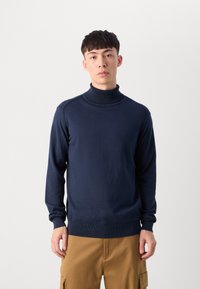 Tamno plavi turtleneck pulover od glatkog pletenog materijala, s dugim rukavima i rebrastim manžetama i rubom. Uparen s svijetlosmeškim cargo hlačama.