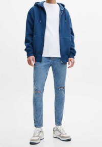 Persona con sudadera azul con cremallera, camiseta blanca, jeans ajustados rasgados de un azul claro, calcetines blancos y zapatillas blancas y grises, de pie sobre un fondo blanco.