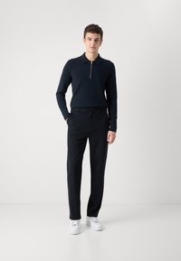 Tengerészkék zipzáras póló, slim fit, fekete nadrággal és fehér tornacipővel kombinálva. Sima anyag, minimalista dizájn.