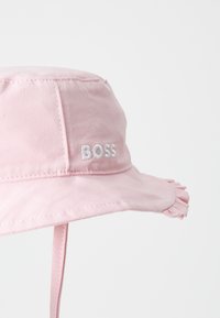 Rosa bucket hat i mjukt tyg, med en volangkant och vit broderad "BOSS"-logotyp på framsidan. Justerbar hakrem ingår.