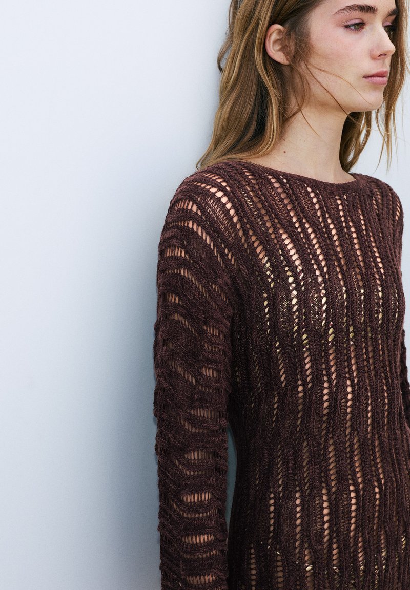 Jeune femme aux longs cheveux portant un pull en maille ajourée brun foncé par-dessus un haut clair, regardant vers la droite sur un fond clair uni.