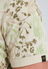 Camicia a maniche corte con una base beige chiaro, caratterizzata da motivi floreali verdi e marroni, tessuto testurizzato e un'etichetta nera con logo sull'orlo.
