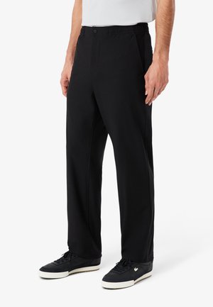 Uomo che indossa pantaloni neri a gamba dritta con vita elastica, abbinati a sneakers nere e una camicia bianca, in piedi contro uno sfondo semplice.