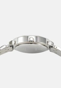 Srebrna ura iz nerjavečega jekla z gladkim okroglim ohišjem, teksturirano krono in svetlim pasom, ki ima vzorec v obliki cikcaka.