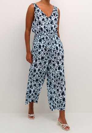 Kukkajumpsuit vaaleansinisessä, jossa tummansiniset kukat, V-kaula-aukko ja elastinen vyötärö. Rennot, lyhyet lahkeet, joissa on sivuaukot liikkumisen helpottamiseksi.