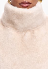 Béžová, měkká směs vlny s vysokým límcem. Textura působí hladce a luxusně, zvyšuje teplo a pohodlí. Jemné, rovnoměrné zbarvení.