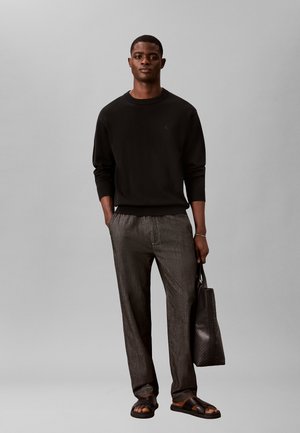 Uomo che indossa una camicia nera a maniche lunghe, pantaloni grigi, sandali, tiene una grande borsa nera e si trova davanti a uno sfondo grigio uniforme.