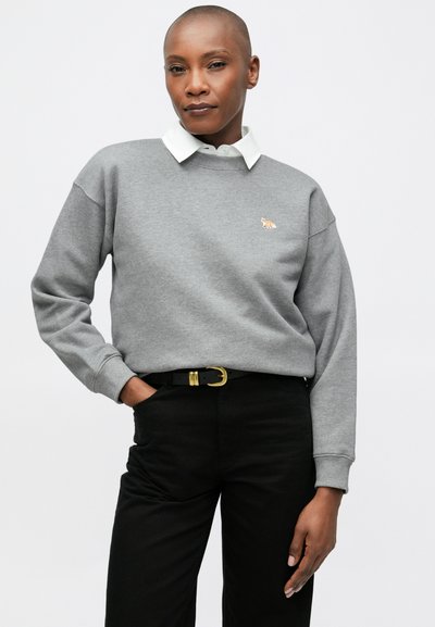 Sudadera gris con un cuello blanco, que presenta un pequeño logo de un zorro naranja. Combinada con pantalones negros y un cinturón negro con accesorios dorados.