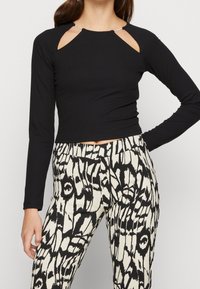 Svart ribbad långärmad topp med unika axelskärningar och silverdetaljer, tillsammans med beige och svarta mönstrade högmidjade byxor.