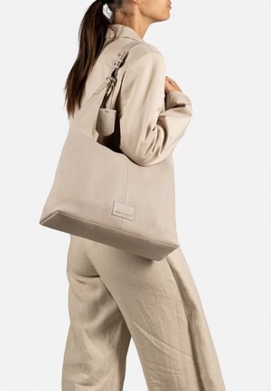 Sac hobo en cuir beige avec une finition texturée, arborant un patch logo bien visible, une bandoulière réglable et une base large et structurée.