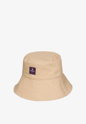 Beżowy kapelusz typu bucket z szerokim przeszywanym rondem oraz małą fioletową kwadratową naszywką z logo "Roxy" z przodu.