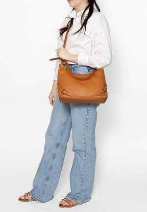 Borsa a spalla in pelle marrone chiaro con una forma arrotondata, caratterizzata da dettagli hardware decorativi e un'unica tracolla, portata da una persona.