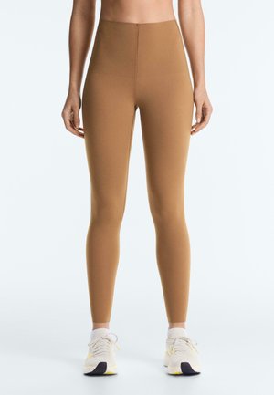 Kobieta w brązowych legginsach z wysokim stanem i białych trampkach, stoi prosto na jasnym, jednolitym tle.