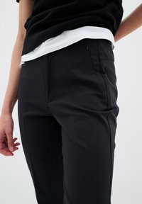Black tailored trousers with a smooth texture, flat front, side pockets, and a sleek design, worn with a layered white and black shirt.