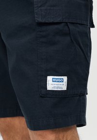 Σκούρο μπλε cargo σορτς με τσέπη με καπάκι που φέρει ένα λευκό ταμπελάκι με την επιγραφή "HUGO" με μπλε γράμματα στον μηρό ενός ατόμου.