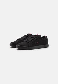 Las zapatillas de lona negras presentan un diseño de bajo corte, cordones en la parte delantera y una suela de goma texturada. Los acentos incluyen detalles de marca en rojo y blanco.