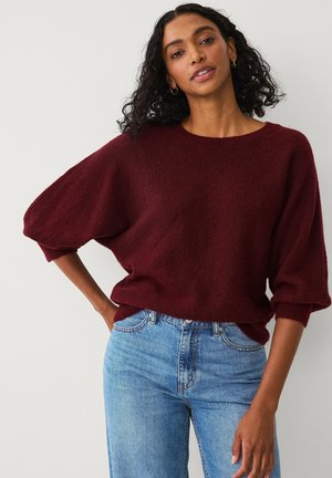 Burgunderfarbener, übergroßer Strickpullover mit weitem Ausschnitt und voluminösen Ärmeln, kombiniert mit hellblauen Denim-Jeans. Weiche Textur und lässige Passform.