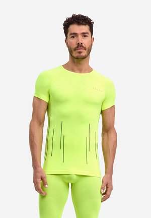 Mann mit kurzen lockigen Haaren und Bart, trägt ein eng anliegendes neon-gelbes Sportshirt und Shorts mit schwarzen vertikalen Streifen auf dem Shirt.