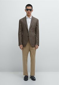 Blazer marrone a trama con rever a punta sopra una camicia bianca, abbinato a pantaloni beige e mocassini neri, indossato da un modello in piedi.