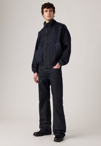 Man met een losse zwarte jas met hoge kraag, bijpassende zwarte broek en zwarte schoenen, staande met één hand in zijn zak tegen een effen achtergrond.