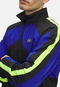 Blauwe en zwarte zipjack met neon groene accenten en een hoge kraag. Bevat een logopatch en een gestructureerde stof. Atletisch ontwerp met lange mouwen.