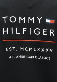Μαύρο ύφασμα με λευκή κεντημένη επιγραφή «Tommy Hilfiger», μια μικρή σημαία με κόκκινο, λευκό και μπλε χρώμα, κόκκινη οριζόντια γραμμή, οι επιγραφές «EST. MCMLXXXV» και «All American Classics».