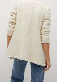 Crème-kleurige blazer van textuurstof, met een losse pasvorm, een rafelige zoom en opgerolde mouwen. Donkerdenim jeans zichtbaar eronder.