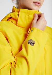 Kobieta poprawiająca kołnierzyk jasnżótej kurtki z małą, teksturowaną łatą na rękawie w pobliżu nadgarstka.