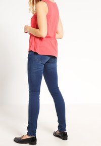 Jeans azules ajustados con una textura suave, combinados con una parte superior sin mangas de color rosa. Se muestra una vista lateral, destacando la forma y las costuras de los jeans.
