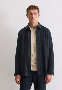 Dunkelmarineblaue Hemdjacke mit Knopfleiste, einer Brusttasche und einem weichen, strukturierten cremefarbenen Pullover darunter. Glatte Stoffoberfläche.