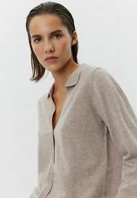 Cardigan beige in maglia morbida con scollo a V, chiusura a bottoni e una trama delicata. Presenta un colletto semplice e maniche lunghe.