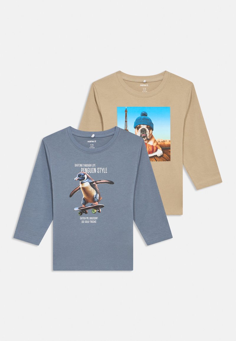 Duas camisetas de manga longa para crianças: uma azul com um pinguim a andar de skate e outra bege com um bulldog a usar um chapéu de malha azul junto à Torre Eiffel.