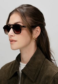 Occhiali da sole con montatura quadrata e motivo tartarugato marrone con lenti sfumate, indossati con una giacca di suede verde scuro e orecchini a cerchio dorati.