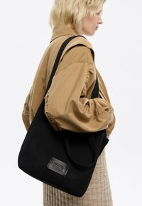 Juodas tinklinis pirkinių maišelis su tekstūruota paviršiumi ir stačiakampio forma, su prekės ženklo odiniu užtrauktuku ir medvilninėmis pečių juostomis.