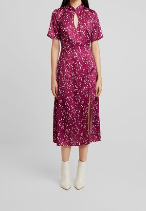 Femme portant une robe rose foncé jusqu'aux genoux avec des taches blanches et violettes, manches courtes, encolure en trou de serrure, fente latérale et bottines blanches.