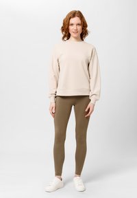 Beżowa bluza z ribbędowymi mankietami i dołem, zestawiona z oliwkowymi legginsami i białymi trampkami. Prosty design, miękka tkanina, dopasowany krój.