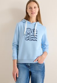 Hoodie bleu clair avec cordon de serrage, présentant un motif de texte ondulé en bleu marine. Tissu doux et lisse avec poignets et ourlet côtelés. Associé à un jean.