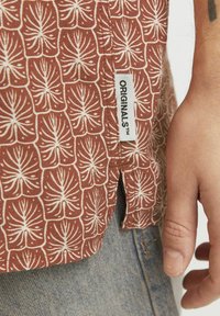 Camisa marrón con estampado de hojas blancas, mangas cortas y una abertura lateral. Se puede ver una etiqueta con "ORIGINALS™".
