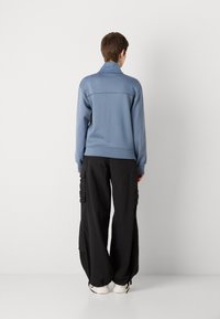Light blue zip-up jacket with a textured finish, worn with loose black cargo pants featuring side pockets. White shoes complete the outfit.
