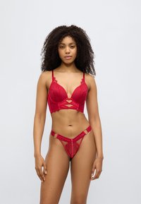Κόκκινο δαντελένιο σουτιέν με βαθύ ντεκολτέ, διακοσμημένο με σχέδιο σταυρωτού τύπου στο κέντρο. Συνοδευτικό κόκκινο δαντελένιο στρινγκ με πλευρικούς κρίκους και διάφανο ύφασμα.