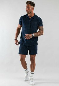 Marinblå pikétröja med en ficka, kombinerad med matchande shorts. Materialet verkar mjukt. Vita sneakers med randiga svarta och vita strumpor.